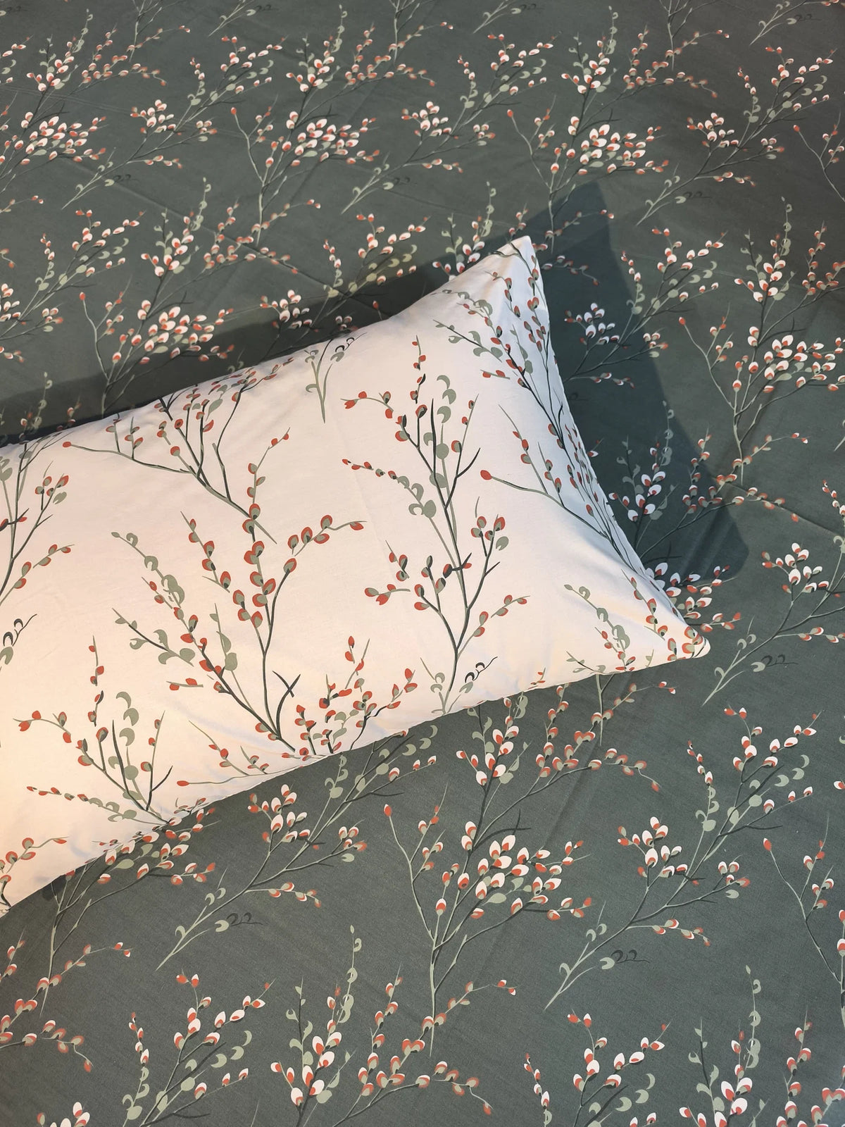 A rectangular decorative pillow with a light cream background and a delicate botanical branch pattern in muted orange and gray-green, resting on a matching dark gray-green patterned bedspread.