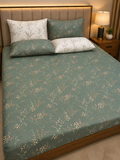 A bed dressed in a sage green floral-patterned sheet set with matching pillowcases and two white pillows with a delicate floral print, shown in a warmly lit bedroom setting.