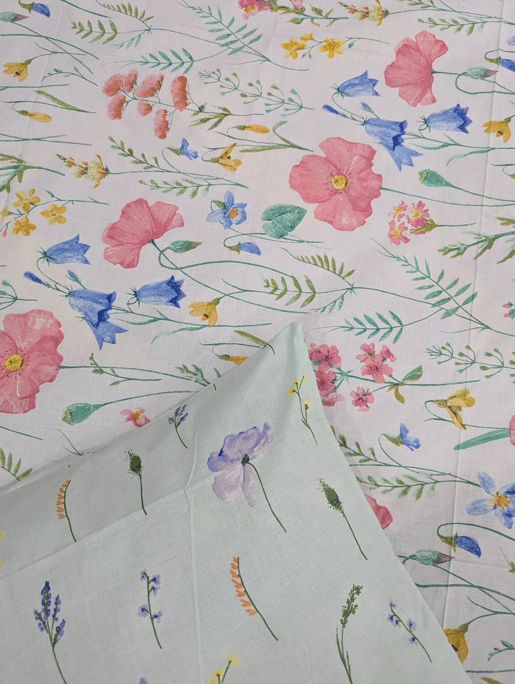 A close-up of a floral fabric with a light pink background, featuring watercolor-style pink, blue, purple, and yellow flowers with green stems and leaves. A folded section of the same fabric is visible in the lower left corner.