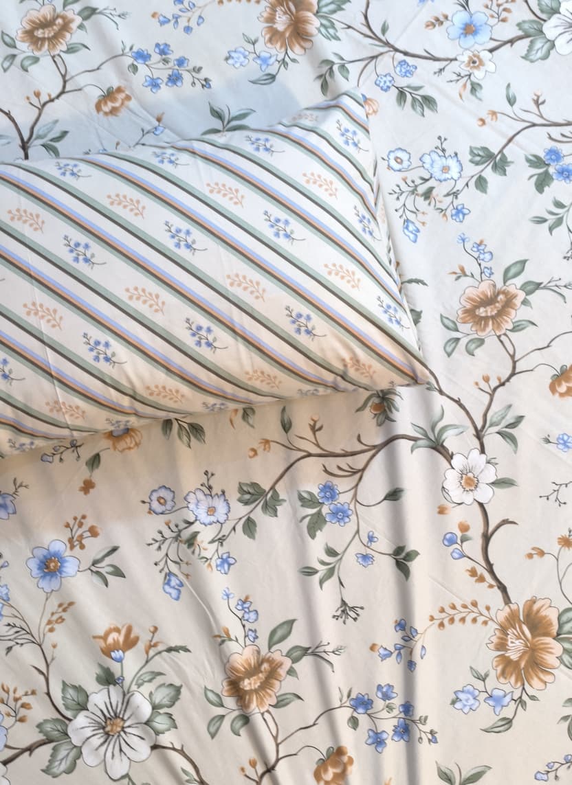 A close-up of a bedding set featuring a cream-colored sheet with a delicate blue, green, and tan floral pattern, paired with a matching pillowcase that has vertical stripes of blue, green, and tan on a cream background.