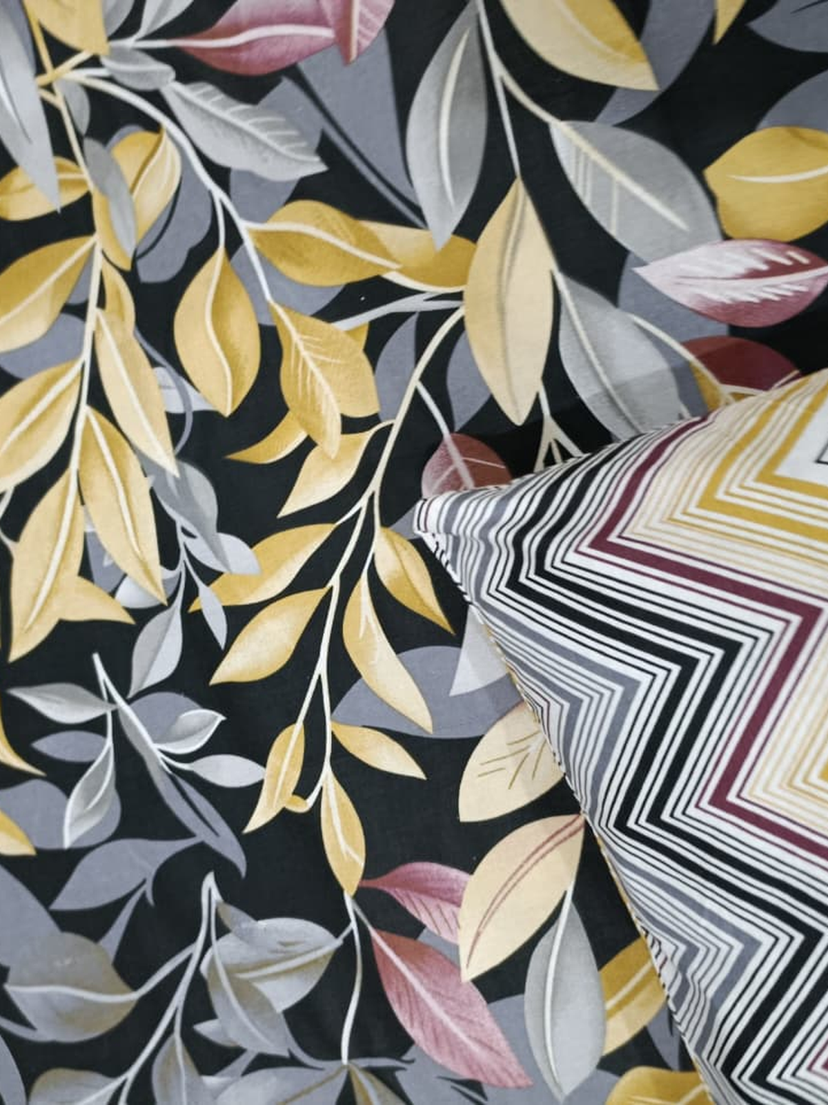 A close-up of a patterned fabric or wallpaper featuring stylized botanical leaves in yellow, gray, and dusty pink on a black background, with a striped chevron-patterned pillow partially visible in the lower right corner.