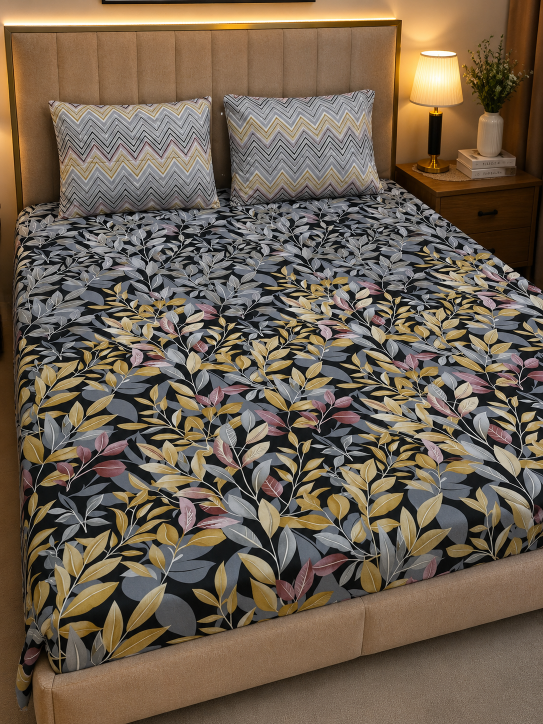 A bed dressed in a dark floral-patterned duvet cover with yellow, gray, and pink leaves, styled with two matching chevron-patterned pillows in a warm bedroom setting.