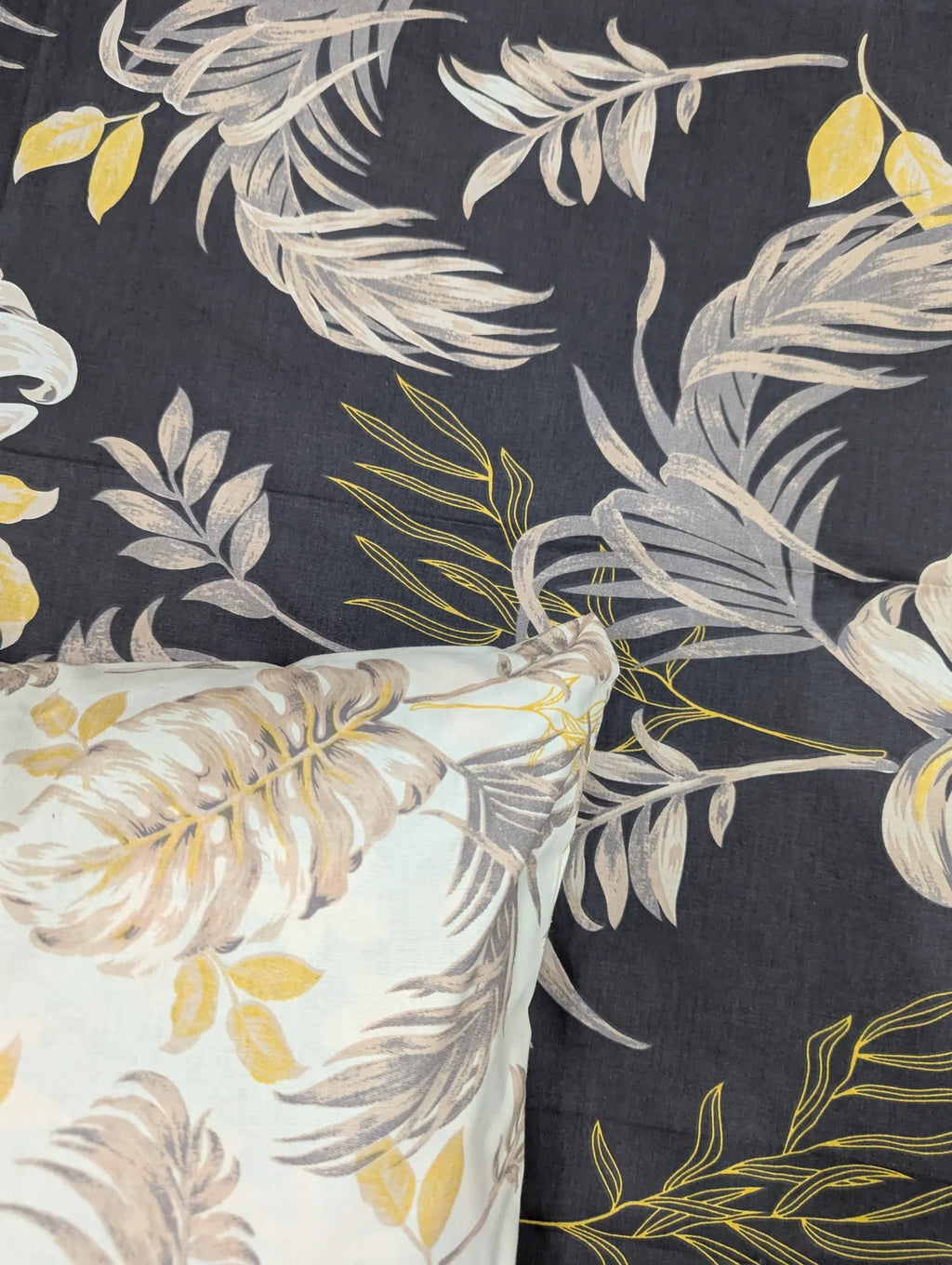 A close-up of a tropical leaf patterned fabric, showing a dark charcoal gray background with large beige and white palm fronds, smaller yellow leaves, and a matching light-colored pillow in the corner.