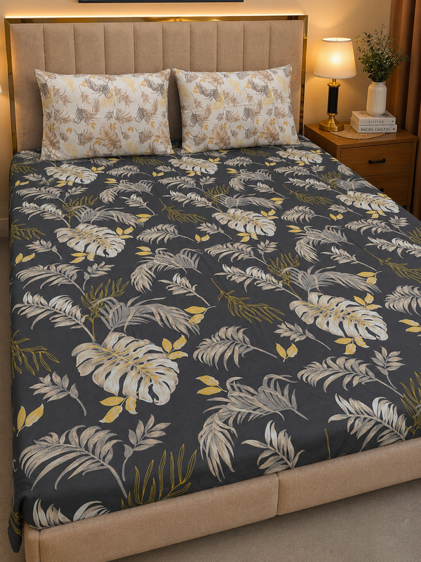 A bed dressed with a dark charcoal gray tropical leaf patterned comforter and matching pillow shams, styled in a warm bedroom setting with a beige upholstered headboard, bedside lamp, and nightstand.
