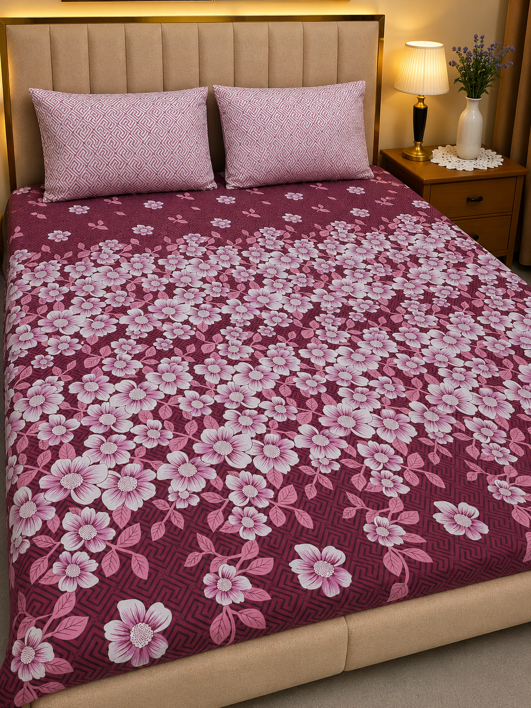 A bed dressed in a dark burgundy floral quilt with pink and white cherry blossom patterns, styled with two matching pink pillows in a bedroom setting.