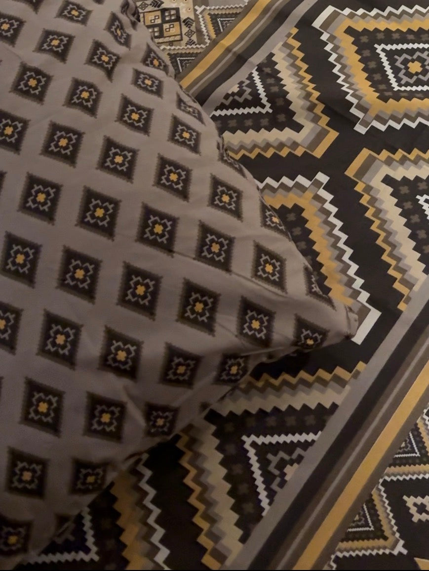 A close-up of a patterned textile, likely bedding or a blanket, featuring a repeating geometric diamond motif in gray, black, white, and yellow-gold, with a matching patterned border visible in the background.