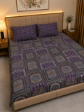 A bed dressed in a purple and blue patterned quilted bedspread with matching pillowcases, set in a styled bedroom with a beige upholstered headboard, bedside tables, and warm ambient lighting.