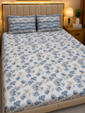 A neatly made bed with a white sheet or duvet cover featuring a blue and gray botanical leaf pattern, paired with two matching patterned pillowcases. The bed is set in a warm, modern bedroom with a beige upholstered headboard, a wooden nightstand, and a lit bedside lamp.