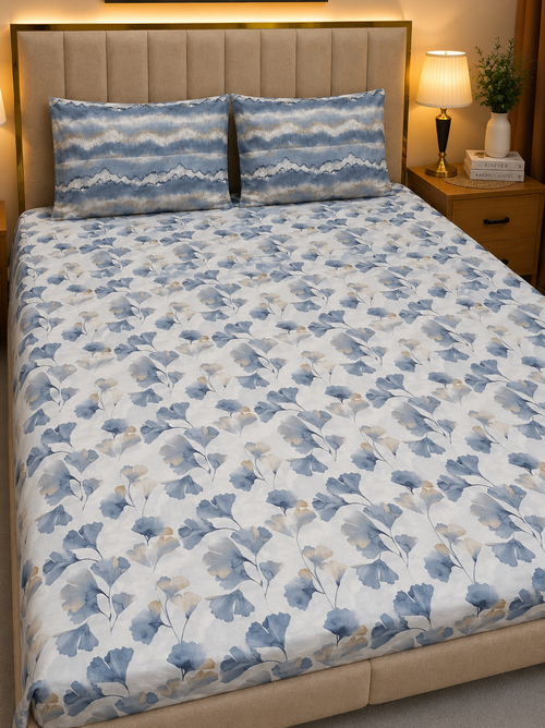 A neatly made bed with a white sheet or duvet cover featuring a blue and gray botanical leaf pattern, paired with two matching patterned pillowcases. The bed is set in a warm, modern bedroom with a beige upholstered headboard, a wooden nightstand, and a lit bedside lamp.