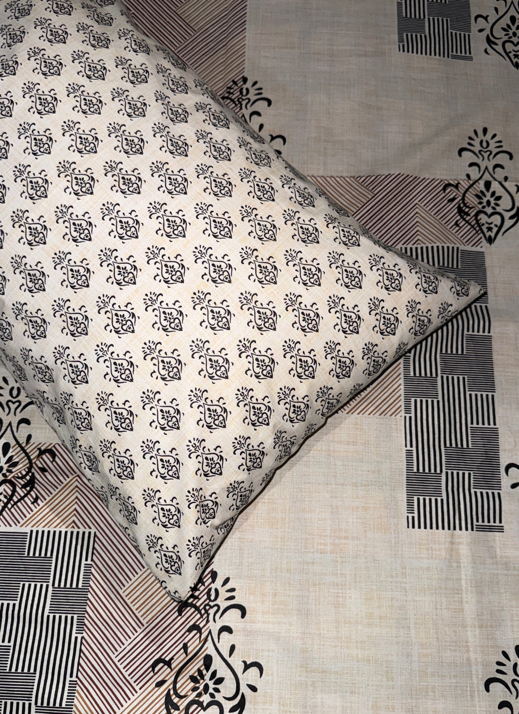A square decorative pillow with a black floral block print on an off-white background, placed diagonally on top of a matching patchwork quilt with black and brown geometric patterns.