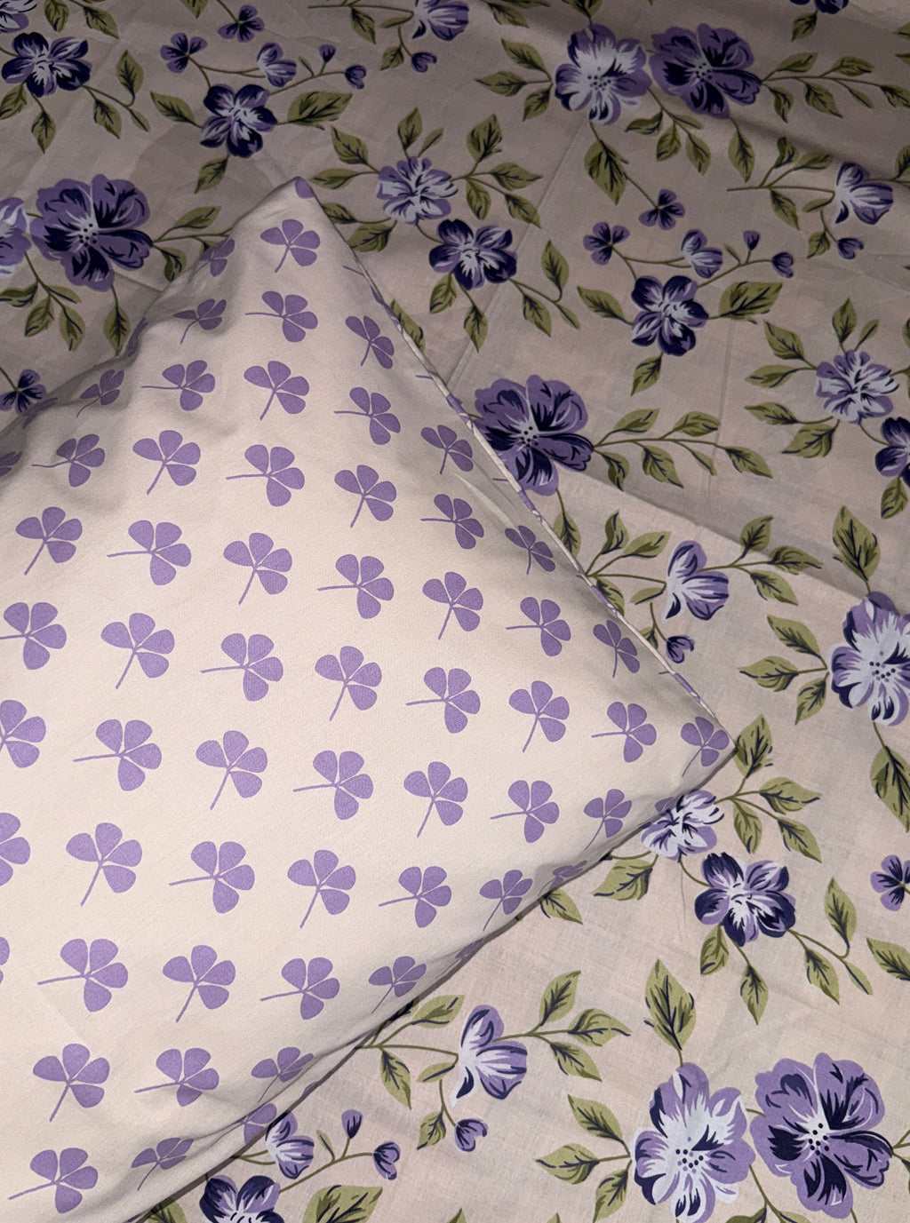 A close-up of a floral fabric product, likely bedding or a textile set, featuring a light beige base with purple flowers and green leaves, layered over a matching sheer mesh sheet with a repeating purple clover pattern.