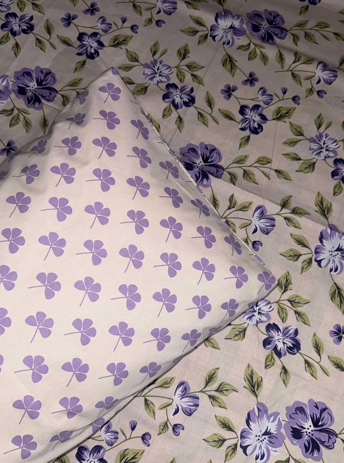 A close-up of a floral fabric product, likely bedding or a textile set, featuring a light beige base with purple flowers and green leaves, layered over a matching sheer mesh sheet with a repeating purple clover pattern.