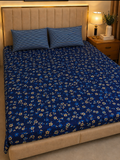 A bed dressed in a dark blue floral-patterned sheet and matching pillowcases, set in a bedroom with a beige upholstered headboard, a wooden nightstand, and a lit lamp.