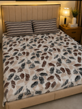A bed dressed in a beige and brown feather-patterned duvet cover with matching striped pillows, shown in a warmly lit bedroom setting next to a bedside lamp.
