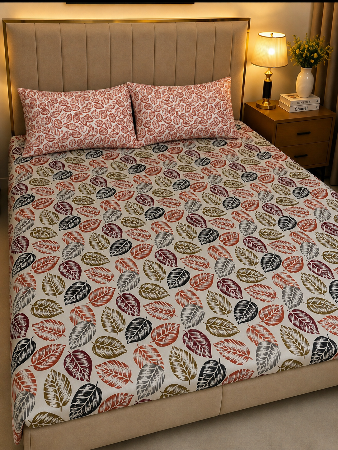 A bed dressed in a patterned duvet cover and matching pillowcases featuring a repeating leaf motif in muted red, olive green, gray, and brown on a cream background, shown in a warmly lit bedroom setting.