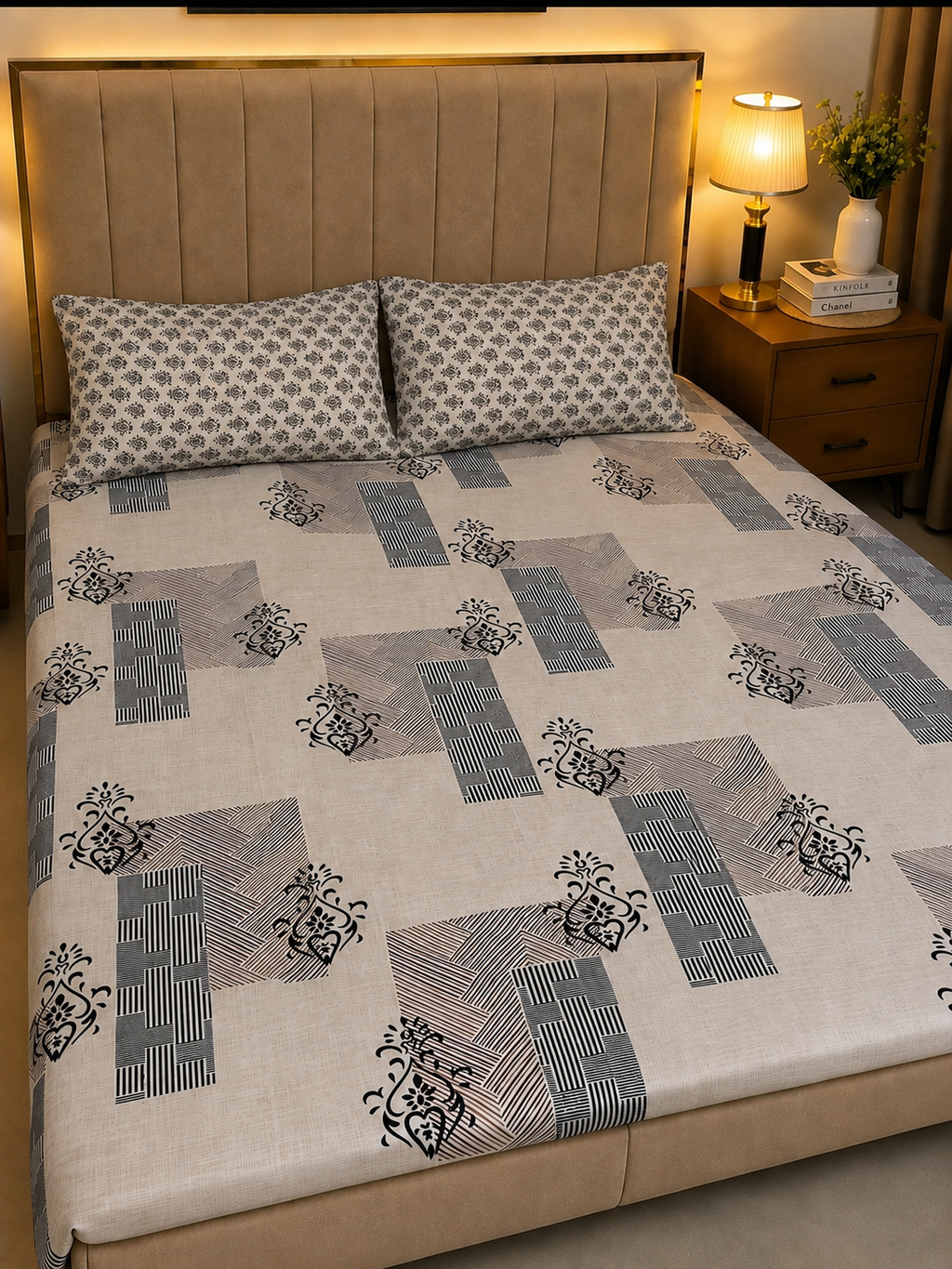 A bed dressed in a patterned bedsheet set with two matching pillowcases, shown in a warmly lit bedroom setting next to a wooden nightstand and lamp.