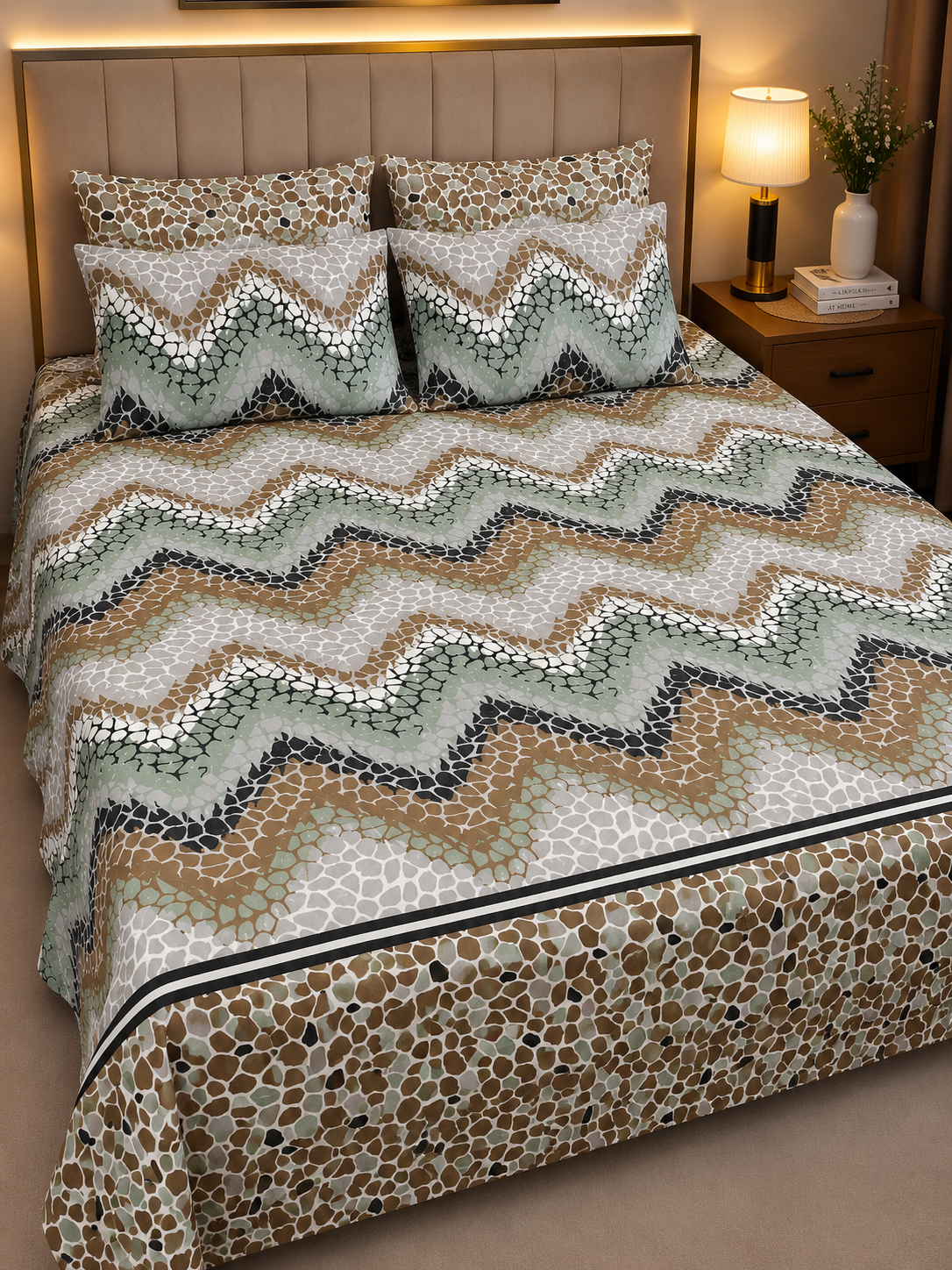 A bed dressed in a patterned bedding set featuring a chevron and animal-print design in beige, brown, sage green, and white, styled in a warm bedroom setting with a headboard, bedside lamp, and plant.