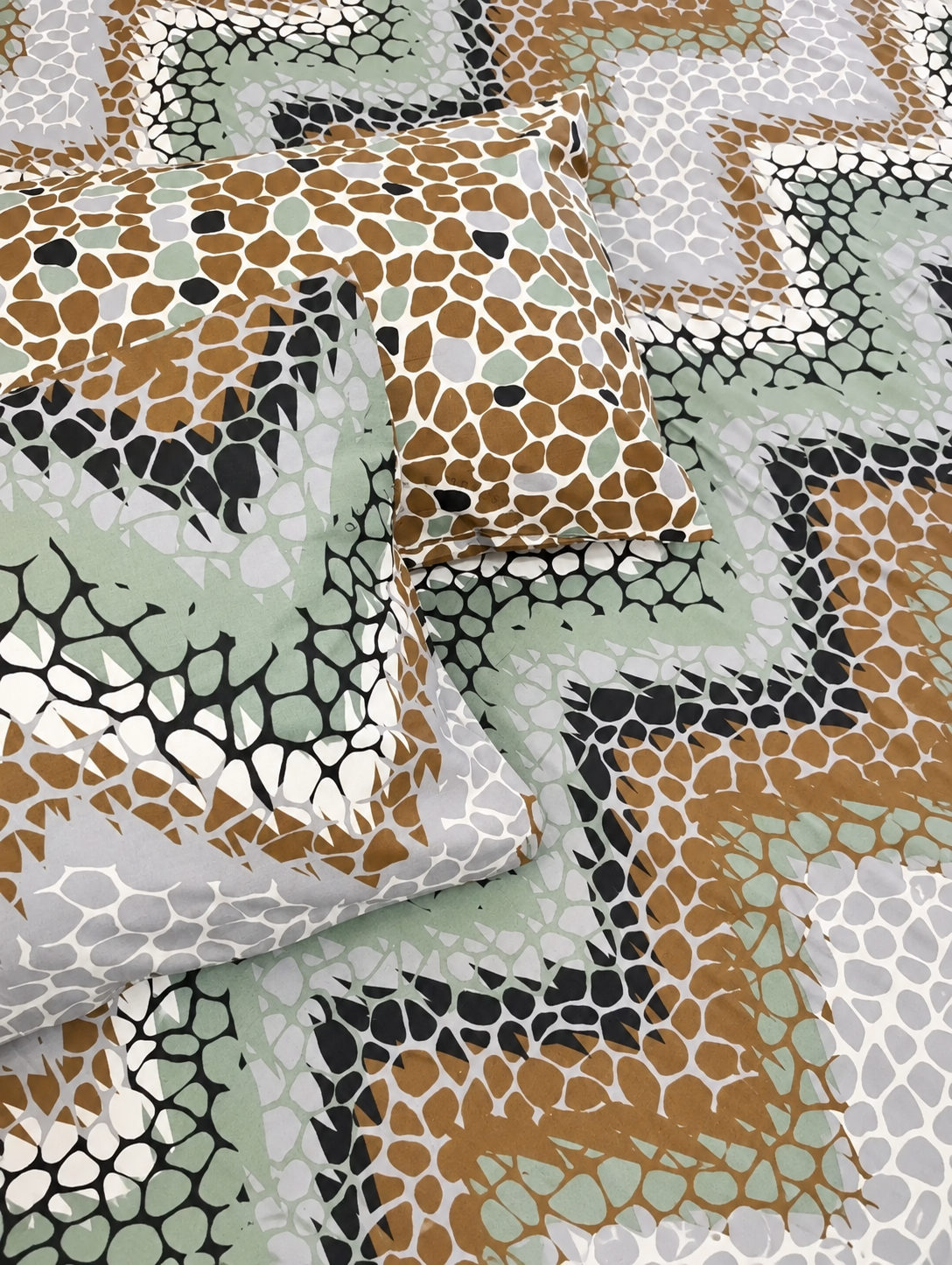 A close-up of patterned bedding or fabric with two matching pillows, featuring a geometric design of tan, sage green, black, and white zigzags overlaid with a cracked reptile-like mesh pattern.
